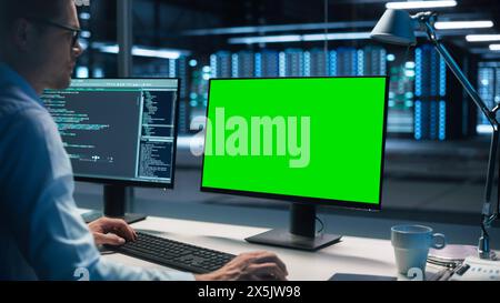 Male Specialist Working on Desktop Computer with Green Screen Mock Up Display in Evening Creative Office. Caucasian Information Specialist working at computer sitting at desk. Over the Shoulder Shot Stock Photo