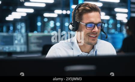 Cheerful Caucasian Male Technical Support Specialist Talks Friendly with a Client While Using Hands-Free Headset at Call Center. Smiling Customer Service Representative in Headset Consulting Clients Stock Photo