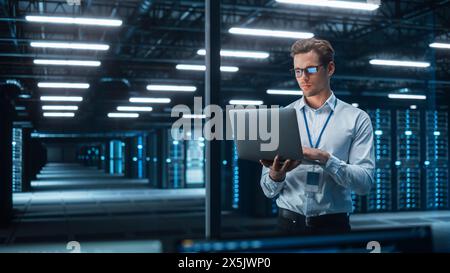 In Technology Research Facility: Chief Engineer Stands in the Middle of the Lab and Uses Laptop Computer. E-Business Digital Entrepreneur Examining Cloud Web Services at the Evening Stock Photo