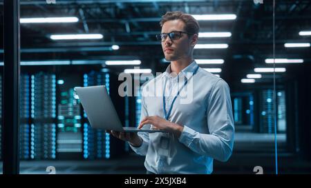 European Male IT Specialist Using Laptop Computer, Standing in Big Warehouse Data Center. System Administrator working with SAAS. E-Business Digital Entrepreneur Examining Cloud Web Services Stock Photo