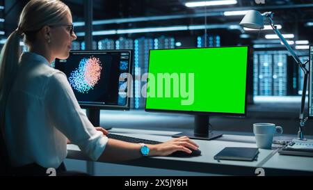 View Over the Shoulder of the IT Technician Working with Neural Network on Her Personal Computer with Green Screen Mock-up. She Works in a Big System Control Data Center Stock Photo