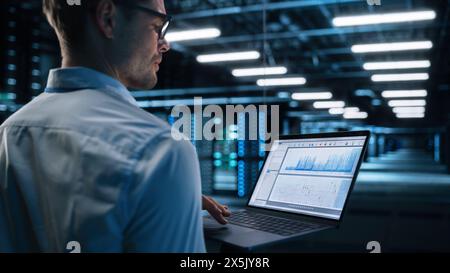 View Over the Shoulder of Male IT Specialist Using Laptop Computer in Data Center, Looking at the Screen. Successful Businessman and e-Business Entrepreneur Working Alone. Innovation Concept Stock Photo