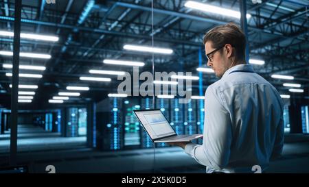 Portrait of IT Specialist Uses Laptop in Data Center. Server Farm Cloud Computing Facility with Male Maintenance Administrator Working. Cyber Security and Network Protection Stock Photo
