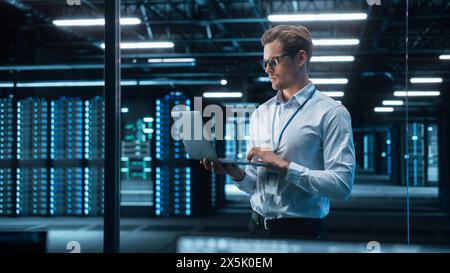 Portrait of IT Specialist Uses Laptop in Data Center. Server Farm Cloud Computing Facility with Male Maintenance Administrator Working. Cyber Security and Network Protection Stock Photo