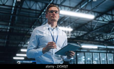 Caucasian Data Center IT Technician Standing at the Server Rack Corridor with a Tablet Computer. He is Visually Inspecting Something while Looking Away. Night Office Concept Stock Photo