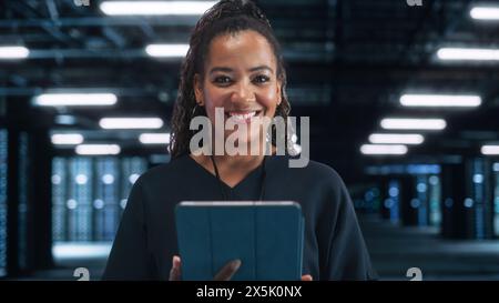 Portrait of the Cheerful Female IT Specialist Using Tablet Computer in Data Center, while Smiling to the Camera. Successful Businesswoman, e-Business Entrepreneur Concept Stock Photo