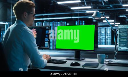 Data Center: Male Office Worker Using Computer with Green Screen Mock Up in Surveillance Center in a Monitoring Office. Specialist Works in Front of Chroma Key Display. Stock Photo