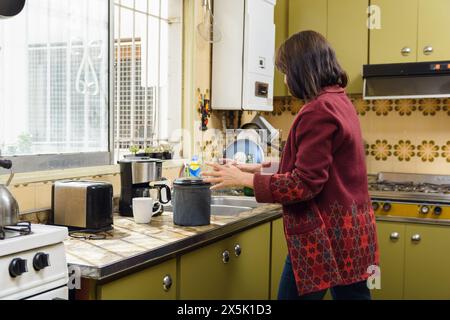 A Latin adult Woman at Home is preparing morning doing household chores, she is wearing red sweater, Kitchen is classic style with green cupboards, wi Stock Photo