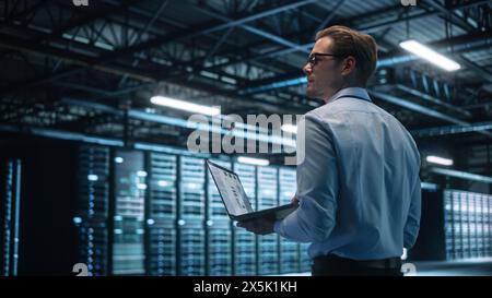 Creative Caucasian Man Using a Laptop Computer in Evening Office. Independent Businessman is Walking Through the Office and Looking Away. Technology Science Breakethrough, Progress, Innovation Concept Stock Photo