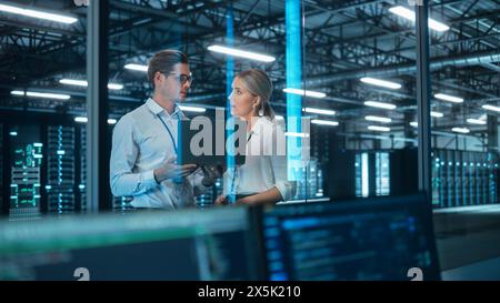 Two IT Engineers Standing in Working Data Center. They Use Laptop while Standing at the Evening Office. Team of Developers Look at Each Other, Talk and Using Computer Stock Photo