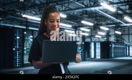 Portrait of Successful African American Female IT Specialist Using Laptop, Standing in Data Center. System Administrator Works on Web Services. Cloud Computing, Server Analytics, Cyber Security Stock Photo