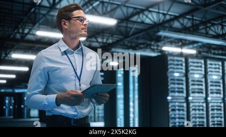 Young Caucasian Male Employee Uses Tablet Computer in System Control Monitoring Center at Night. Evening Office In the Background. Cyber Security and Network Protection Concept Stock Photo
