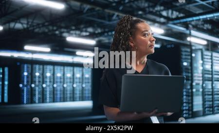 Female Mature Multiracial e-Business Entrepreneur Look Away and Holding Laptop at Data Center Server Evening Room. Technology Web Services Cloud Computing and SAAS Concept Stock Photo