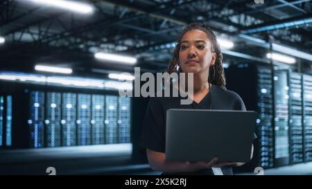 Mixed RaceFemale IT Specialist Uses Laptop Computer in Data Center. Server Farm Cloud Computing Facility with Maintenance Administrator Working. Cyber Security Engineer Working in Network Protection Stock Photo