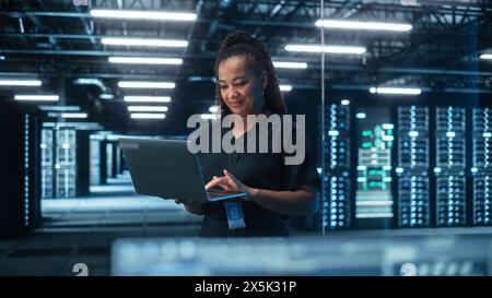 Portrait of Multiracial Mature Female IT Specialist Using Laptop, Standing in Data Center. Infrastructure Architect Works on Web Services. Cloud Computing, Server Analytics, Cyber Security Maintenance Stock Photo