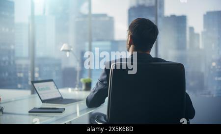 Relaxed Businessman in a Suit Sitting at a Desk in Modern Office, Using Laptop Computer, Looking at Window City View. Successful Finance Manager Stretching at His Desk while Planning Work Projects. Stock Photo