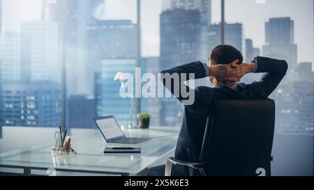 Relaxed Businessman in a Suit Sitting at a Desk in Modern Office, Using Laptop Computer, Looking at Window City View. Successful Finance Manager Stretching at His Desk while Planning Work Projects. Stock Photo