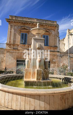 Alberobello, Italy. Piazza water fountain with Roman urns and face mosaic, limestone building and blue shutters Stock Photo