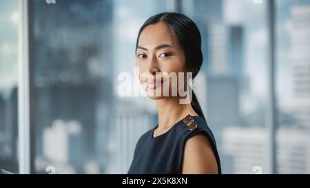 Beautiful Portrait of an Asian Businesswoman in Stylish Black Dress Posing Next to Window in Big City Office with Skyscrapers. Confident Female CEO Smiling. Successful Diverse Business Manager. Stock Photo