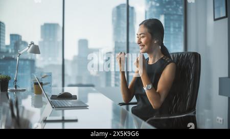 Excited Businesswoman in Stylish Dress Sitting at a Desk in Modern Office, Using Laptop Computer. Successful Manager Ecstatic About Winning a Real Estate Investment Deal for Corporate Business. Stock Photo