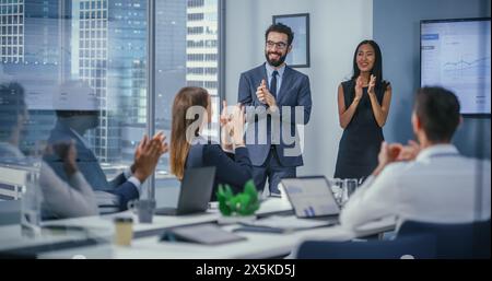 Office Conference Room Meeting: Male Executive and Asian Female Top Manager Present e-Commerce Startup Statistics on Wall TV to a Group of Investors. Everybody Applaud and Celebrates Great Results Stock Photo