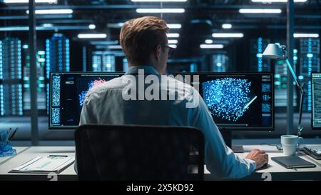 Software Developer Works with AI Analysis on his Computer with Two Monitors. Developer Coding while Looking at the Display of Computer. Modern Evening Dark Office. Back view. Stock Photo