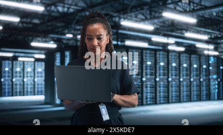 Successful Female IT Specialist Using Laptop Computer, while Standing at Big Warehouse Data Center. System Administrator working with Computing SAAS, Cloud Services. E-Business Digital Entrepreneur Stock Photo