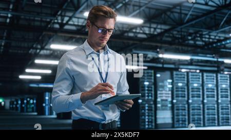 Handsome IT Specialist Using Tablet Computer in Data Center, Looking at the Screen. Succesful Businessman and e-Business Entrepreneur Overlooking Server Farm Cloud Computing Facility. Stock Photo