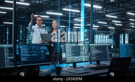 Successful Corporate Top CEO's Talking, Using Laptop Computer Standing in Big City Office at Night. Two Successful Managers Discuss Environmental, Social, Corporate Governance for e-Commerce Project Stock Photo