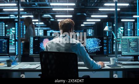 In the Futuristic Laboratory or Office Male Scientists Working at the Two Monitors. Developer Writing Code at the Evening. Computer Engineer Create Neural Network at His Workstation Stock Photo