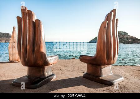 a pair of hand-shaped wooden chairs installed on the beach of Gumusluk, a small fishing village near Bodrum, allows tourists to relax at sunset Stock Photo