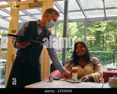 Waiter in face masks serving latte to smiling woman in cafe Stock Photo