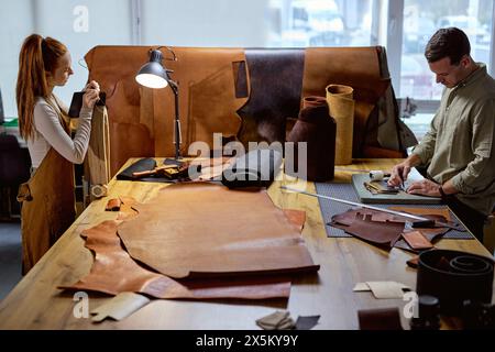 ginger woman in apron using stitching clamp at work, her colleague making holes on leather material with awl. side view shot Stock Photo