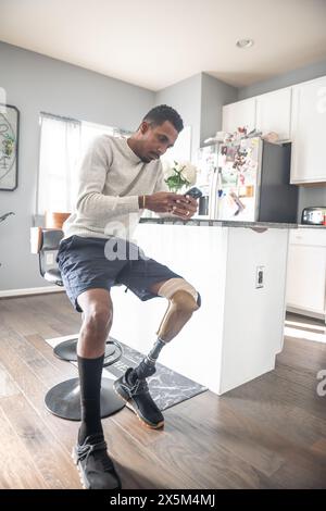 Man with prosthetic leg using smart phone in kitchen Stock Photo