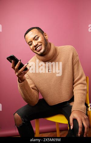 Studio shot of smiling queer man sitting on chair with smart phone Stock Photo