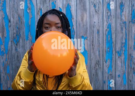 UK, South Yorkshire, Portrait of woman with braided hair blowing orange balloon Stock Photo