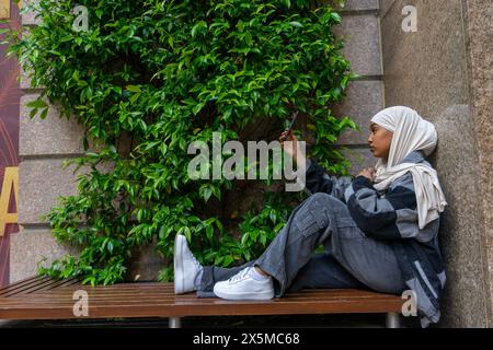 Young woman wearing hijab sitting on bench, taking selfie Stock Photo