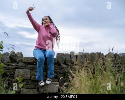 Teenage (16-17) girl sitting on stone wall taking selfies Stock Photo