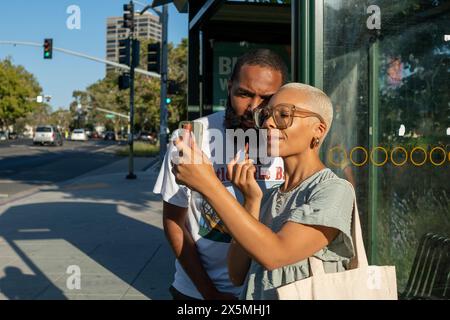 Couple waiting at bus stop and woman applying lip gloss Stock Photo