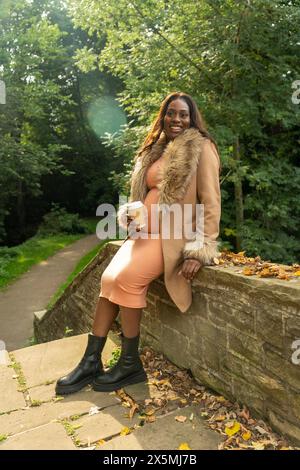 Smiling pregnant woman in coat sitting on wall in park in autumn Stock Photo
