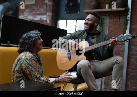 Young man playing acoustic guitar in office lounge, woman laughing Stock Photo