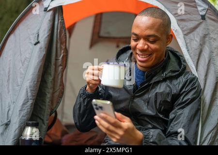 Smiling man using smart phone outside of tent and holding metal mug Stock Photo