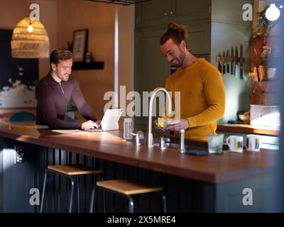Man washing dishes while boyfriend using digital tablet at home kitchen Stock Photo
