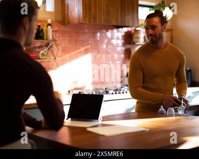 Man washing dishes while boyfriend using digital tablet at home kitchen Stock Photo