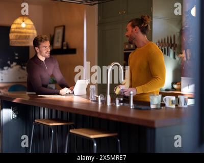 Man washing dishes while boyfriend using digital tablet at home kitchen Stock Photo