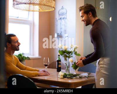 Gay man arranging rose flowers in vase while boyfriend drinking wine Stock Photo