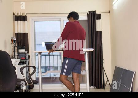 A young man stands at his home office desk, engrossed in work on his laptop. This image captures the essence of working remotely Stock Photo