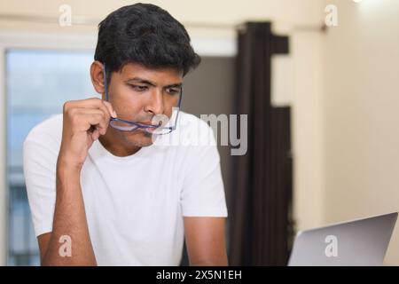A concentrated young Indian man wearing eyeglasses works diligently on his laptop computer at his home office. Stock Photo