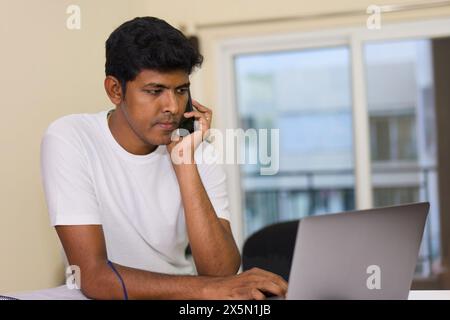 A focused young Indian man juggles tasks seamlessly, using a laptop and talking on a phone in his comfortable home office. Stock Photo