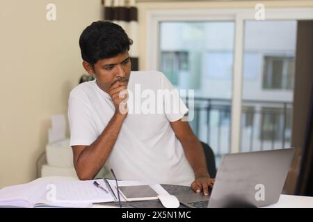 A young Indian man, deep in thought, sits at his home office desk with a laptop computer in front of him. Stock Photo